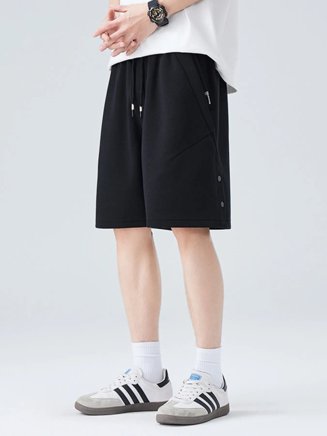 A person wearing black drawstring shorts, a white top, white socks, and white sneakers with black stripes, standing against a light gray studio background.