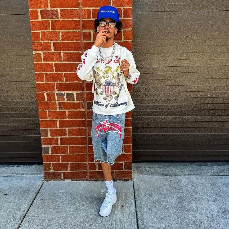 Person wearing a white graphic t-shirt, blue cap, and denim shorts standing against a brick wall.