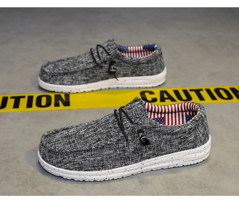 A pair of gray heathered knit slip-on shoes with white soles, displayed on a gray concrete floor with a yellow caution tape prop.