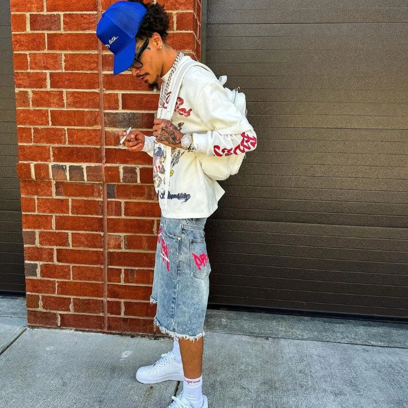 Person wearing a white jacket with red text, denim shorts, and a blue cap leaning against a brick wall.