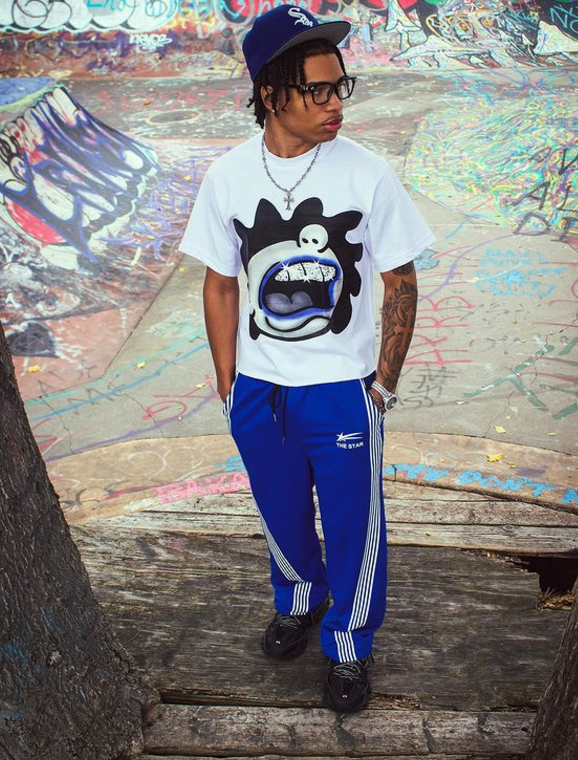 Person wearing a white t-shirt with a graphic design and blue pants standing against a graffiti-covered wall.