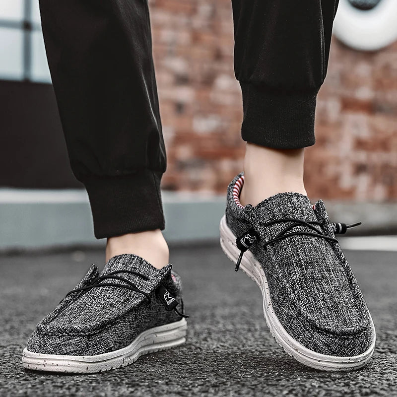 A person wearing black jogger pants and gray knit slip-on sneakers with black laces, standing on a paved surface with a blurred brick wall in the background.