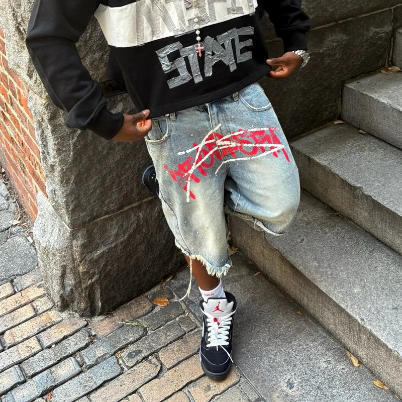 Person wearing a black and white hoodie with 'STATE' text, denim shorts with red paint, and black sneakers on stone steps.